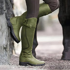 Side view of khaki green suede knee-high boot with buckle strap, worn against a stone wall