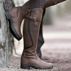 Weathered brown leather knee-high boot with buckle strap shown in profile against a stone wall