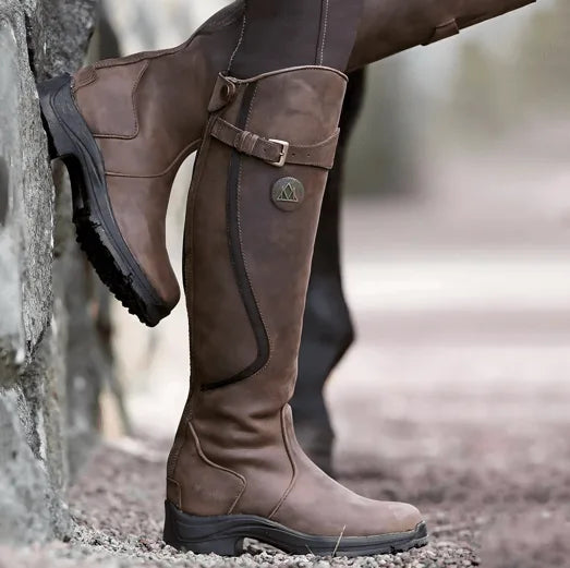 Weathered brown leather knee-high boot with buckle strap shown in profile against a stone wall