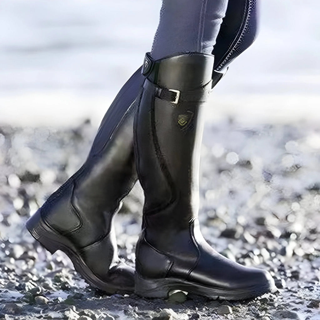 Close-up profile of glossy black leather knee-high boot with buckle strap on a pebbled path