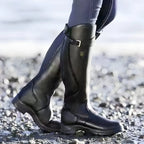 Close-up profile of glossy black leather knee-high boot with buckle strap on a pebbled path