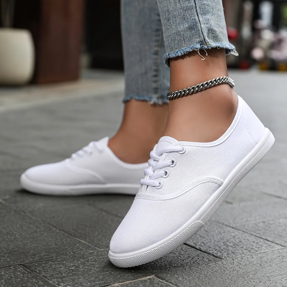 Close-up side view of white canvas lace-up trainers worn with cropped jeans and a silver chain anklet on a stone pavement.