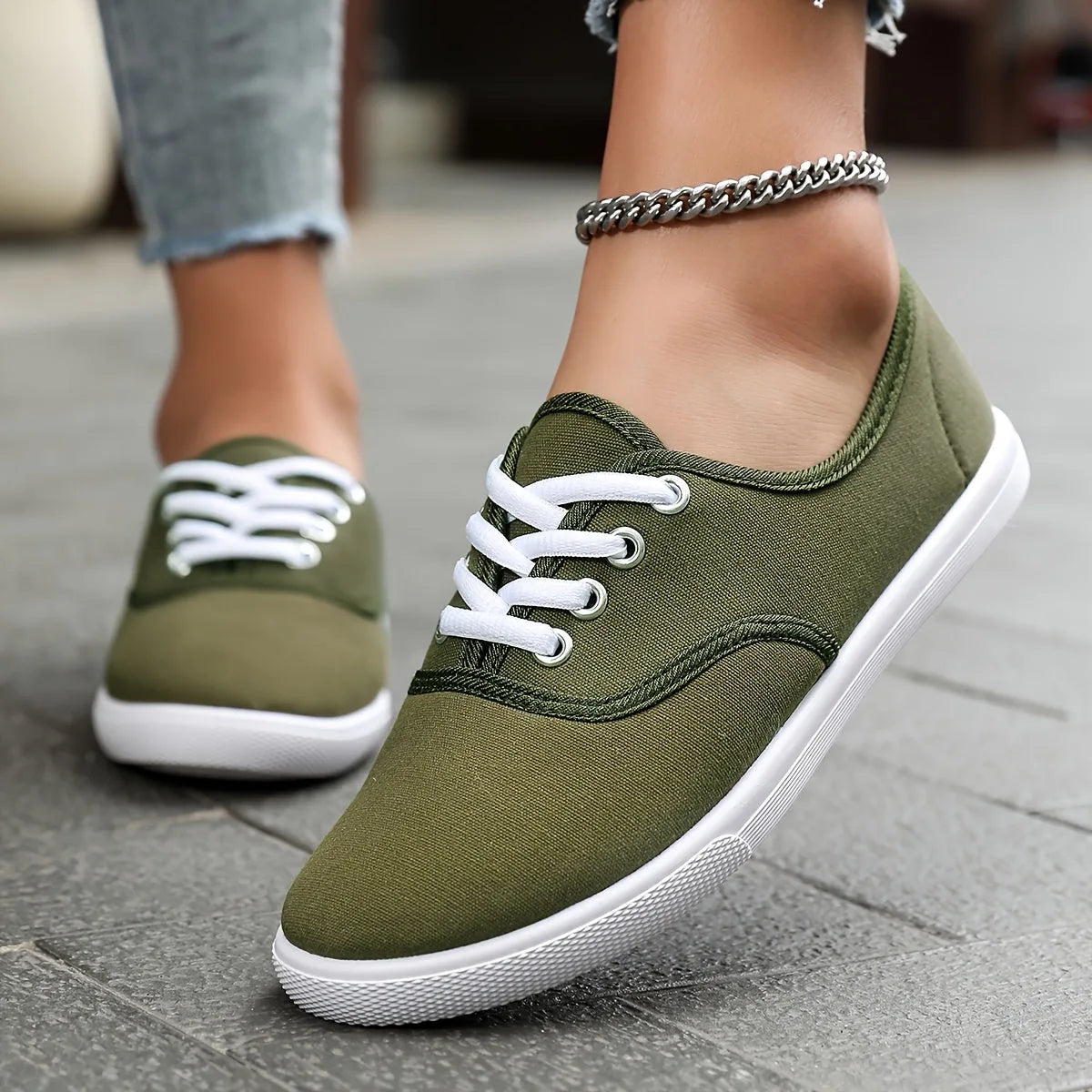 Khaki green canvas lace-up trainers with white laces and soles, close-up side angle on a stone pathway.