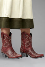 Close-up front view of red leather ankle-high cowboy boots peeking under a cream and olive skirt hem