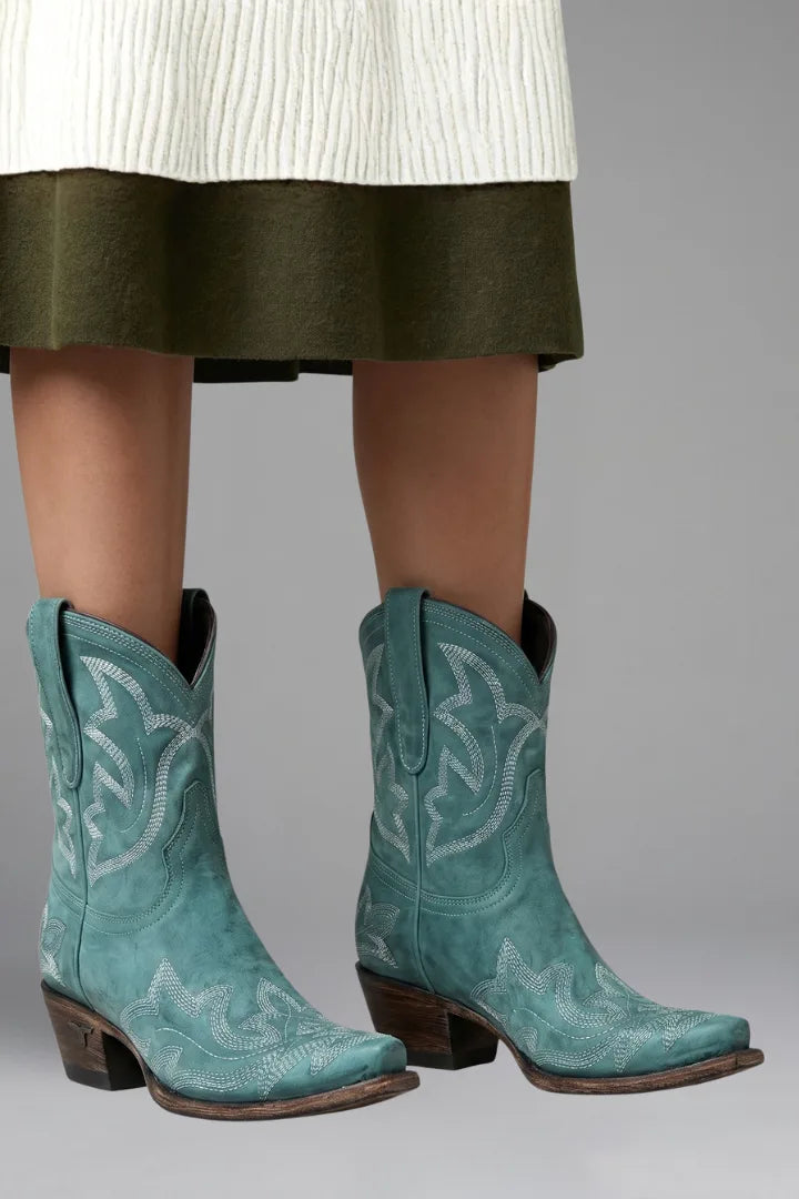 Front view of turquoise teal suede-look cowboy boots with cream embroidery worn beneath a knit hemline