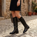 Woman walking down a cobbled street wearing black knee-high leather boots with a black skirt and tote bag