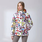 Front-facing shot of the pink floral hooded jacket paired with grey trousers against a snowy backdrop
