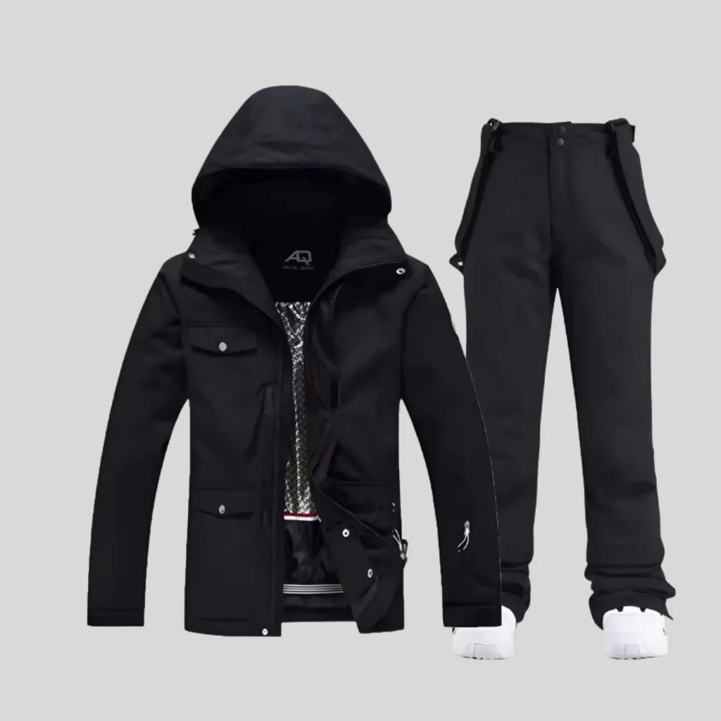 Black two-piece ski set with hooded jacket and bib-style trousers, worn with white trainers