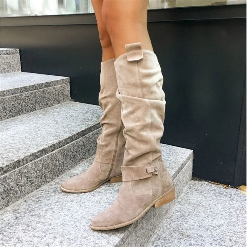Side view of beige suede knee-high slouch boots with buckle strap detail, worn on stone steps