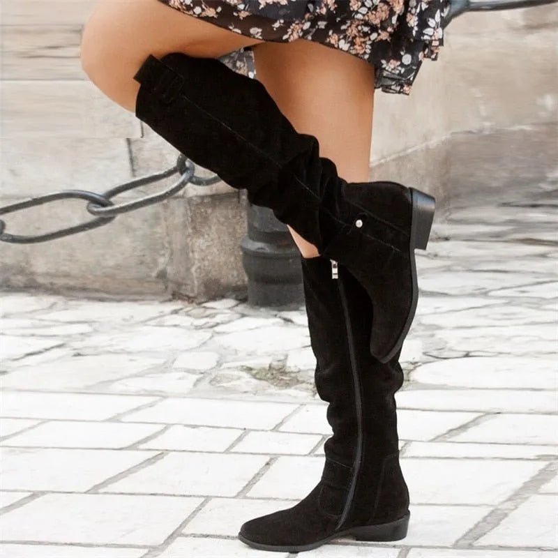 Black suede knee-high slouch boots worn with a floral skirt, close-up angled view on cobblestones