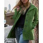 Green textured coat with turndown collar and button-front, worn over a black top with jeans, shown against a brick wall